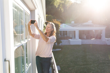 Happy senior caucasian woman taking selfies, standing on sunny balcony