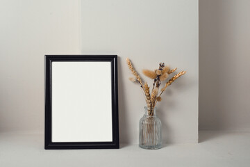 Still life with a black frame. Next to it is a glass vase with dried herbs. Light background.
