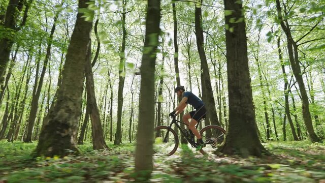 A cyclist on an MTB bike is riding through the forest at high speed. The camera is moving parallel to it. The sun is shining through the trees. He is wearing cycling gear. 4K 