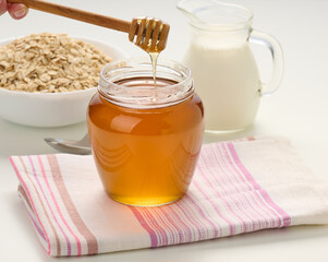 honey in a glass transparent jar and a wooden stick on a white table, behind a decanter with milk...