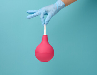 a hand in a blue medical glove holds a pink rubber enema on a blue background