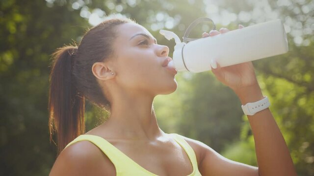 Close up portrait of young african american sporty lady drinking water from sports bottle in public park, tracking shot