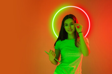 Bright portrait of sweet young woman isolated on orange studio background in multicolored neon light with geometric luminescent shape cirlce