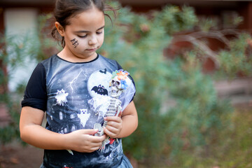 Little caucasian child disguised as a witch with toy skeleton in her arms. She is outdoors. Space for text.