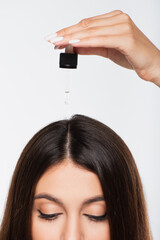 cropped view of young woman with shiny hair applying treatment oil on top of head isolated on grey