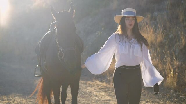 Young slim woman in white shirt and straw hat walking in slow motion holding horse bridle. Portrait of confident gorgeous Caucasian rider with Russian Happiness tattoo strolling with graceful animal