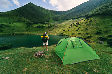 A male tourist with a backpack and a rubber mat stands near a tent by the lake. A young hiker at the foot of the green mountains. Travel, vacation, tourism.