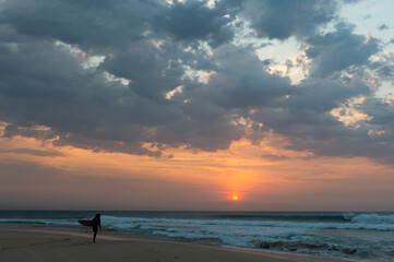 The sun rises over the ocean as a surfer walks along the beach