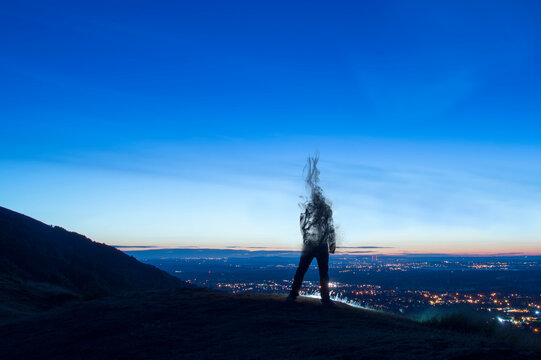 A Silhouette Of A Man Disappearing And Turning Into Smoke. Standing On A Hill. Looking Out On City Lights Just Before Sunrise.