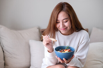 A young asian woman eating healthy blueberry smoothie bowl with mixed fruits topping on sofa at home