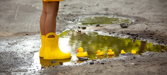 Little girl in a yellow dress and yellow rubber boots lets a family of yellow ducks in a puddle. Autumn and spring consept. Summer rain. Spring Girl under a rainbow umbrella