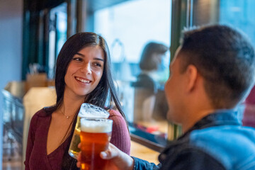 Young couple in love flirting and toasting with glasses of beer, young people having fun at pub, smiling brunette woman