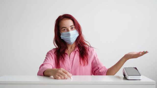 Woman Administrator Of A Sales Area In A Pink Shirt And A Medical Mask On Her Face Stands Behind The Reception And Invites Clients To The Store On A White Background. Small Business Concept.