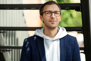 Portrait of casual man in eyeglasses wearing trendy hoodie and blazer standing outside office...