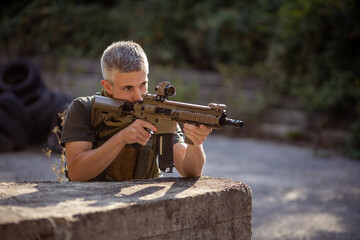 A man kneels behind the cover with an airsoft assault rifle in military uniform