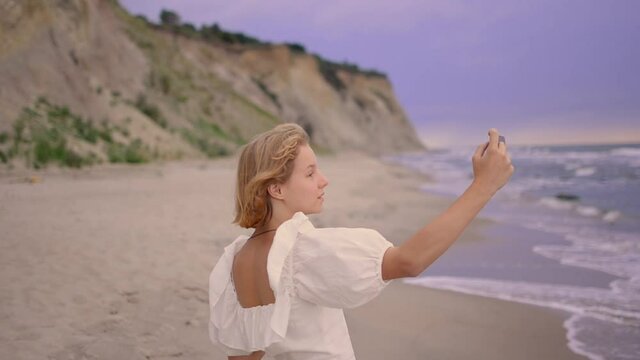 Close up of cheerful young blond girl in white dress taking a selfie at the beach.Selfie girl taking picture on travel holiday in vacation destination. Blond girl holding mobile photo to take self-por