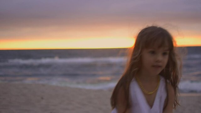 Adorable happy little girl in white dress 4-5 years is dancing and playing on white beach at sunset. Cute kid dance on the seashore.