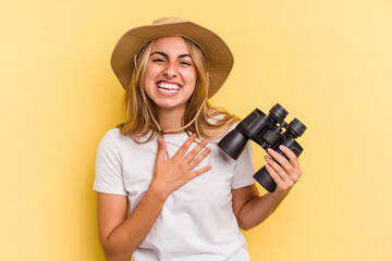 Young caucasian woman holding binoculars isolated on yellow background  laughs out loudly keeping hand on chest.