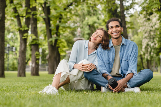 Young mixed-race couple boyfriend and girlfriend students spending time together on romantic date sitting on green lawn grass in city park walking outdoors. Love and relationship concept