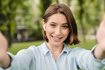 Smiling young caucasian woman girl student taking photo selfie on smart phone outdoors in city park listening to the music in earbuds.