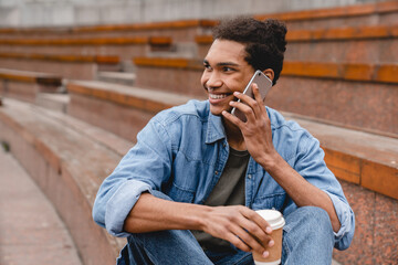 Close up young african student man talking on smart phone with perfect connection sitting on a bench in urban area. Young freelancer relaxing communicating with friends on cellphone outdoors.