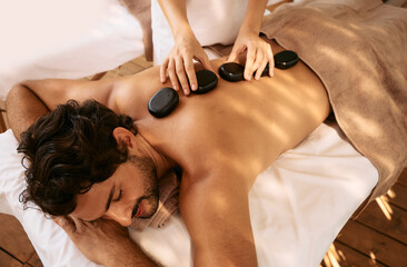 Handsome man at spa resort receives hot stone massage. Hot stone massage therapy using smooth,...