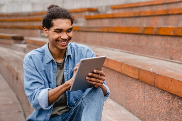 Young african man student freelancer using digital tablet reading e-book, working remotely, surfing on Internet, social media, checking e-mails, e-learning, e-banking online in urban area