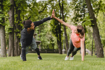 Fit healthy young male and female athletes boyfriend and girlfriend friends couple standing in plank position stretching warming-up before training workout together outdoors in city park.