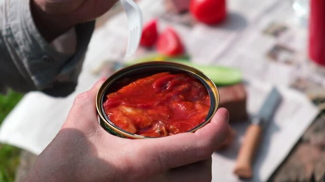 A man takes out canned fish in tomato sauce with a fork. Picnic in nature. In the background an impromptu table with vegetables and bread. Fast food, delicious snack.
