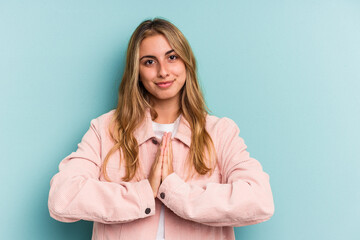 Young caucasian blonde woman isolated on blue background  praying, showing devotion, religious person looking for divine inspiration.