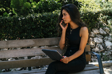 Fototapeta premium a brunette girl sitting on a bench using a tablet and talking on the phone