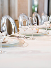 Table serves for banquet. Transparent wine glasses, plates with napkins and shiny cutlery on white table cloth at sunlight.