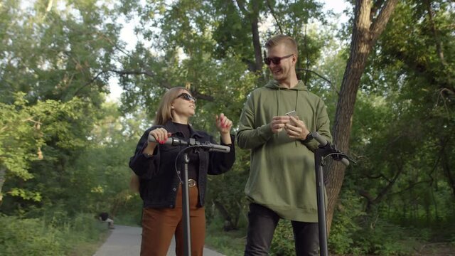 Happy couple with glasses take selfies on their phone and ride electric scooters. Man and woman of caucasian appearance. Concept of traveling on electric scooters. Wanderlust