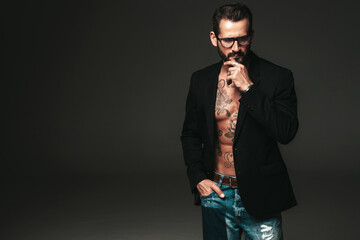 Fototapeta premium Portrait of handsome confident stylish hipster lambersexual model. Sexy modern man dressed in black jacket. Naked torso with tattoos.Fashion male posing in studio on dark background. In spectacles
