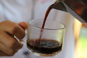 A man pouring fresh hot coffee from geyser coffee maker into glass cup.