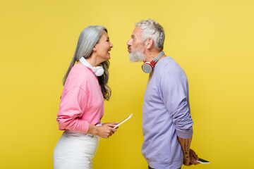 side view of funny multiethnic and middle aged couple in wireless headphones looking at each other and grimacing isolated on yellow - Powered by Adobe