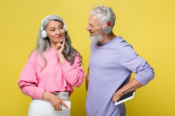 cheerful middle aged and multiethnic couple in wireless headphones looking at each other and...