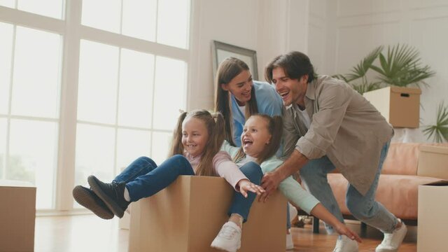 Funny active family playing on moving day, excited mom and dad pushing cardboard box with cute little daughters