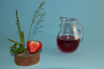 Strawberry on a piece of wood with leaves, a jug with fresh juice in the background. Everything is on a light blue background.