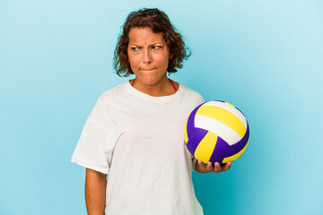 Middle age latin woman playing volleyball isolated on blue background confused, feels doubtful and unsure.