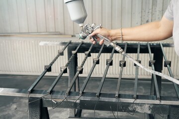 Woman worker paints forged balusters in black color with spray gun on rack support in contemporary...
