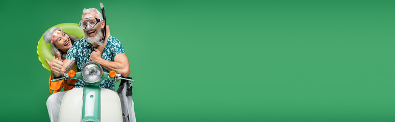 cheerful middle aged asian woman hugging husband in goggles showing thumb up while riding moped isolated on green, banner © LIGHTFIELD STUDIOS