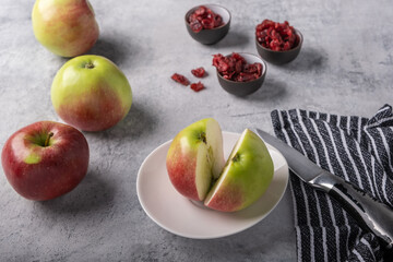 Freshly cut appetizing red apple on a plate, next to whole large beautiful apples, a knife on a napkin, bowls with dried cranberries on a white concrete background.