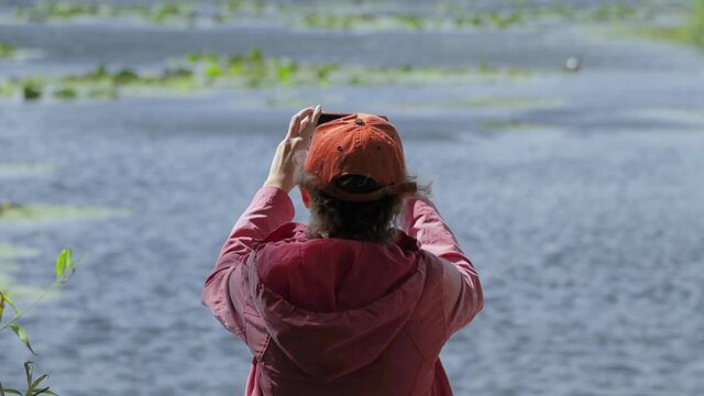 A girl in a red jacket and a baseball cap is sitting on the shore and taking pictures of the river on her smartphone. Shooting from the back. Soft focus