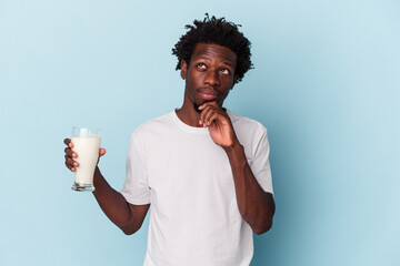 Young african american man holding a glass of milk isolated on blue background looking sideways with doubtful and skeptical expression.