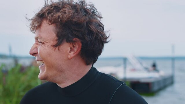 Wake surfer interview. Portrait of the talking man in black wetsuit standing outdoor with lake and boat on the background