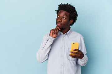 Young african american man holding mobile phone isolated on blue background looking sideways with doubtful and skeptical expression.