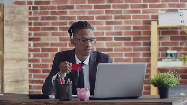 Black man in glasses and a suit enters the bank card data into a laptop at the office table in office.