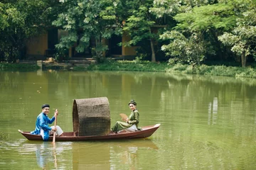 Foto op Canvas Smiling just married Vietnamese couple in traditional dresses and headwear rowing boat in small pond © DragonImages