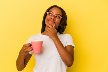 Young african american woman holding a mug isolated on yellow background  looking sideways with doubtful and skeptical expression.